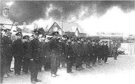 Treowen Road, August 1940. Tired firemen at roll call by Pennar School. Behind them, acrid smoke pours from the burning oil tanks. 
